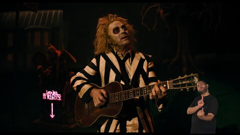 Beetlejuice toca um violão e olha para algo fora de quadro. Uma lápide atrás dele diz: “Aqui jaz Betelgeuse.” O intérprete de Libras, Andrey Baptista, está no canto inferior direito e sinaliza "um".