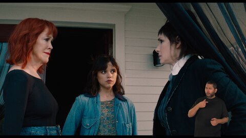 Delia Deetz e Astrid Deetz olham para Lydia Deetz, que está segurando uma cortina preta translúcida aberta.O intérprete de Libras, Andrey Baptista, está no canto inferior direito e sinaliza "aquilo".