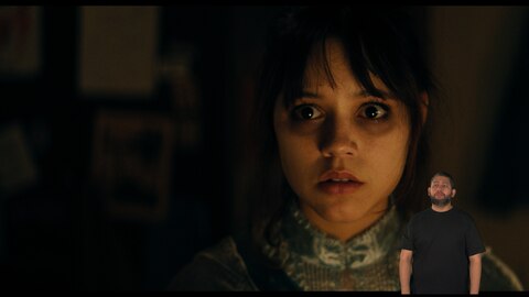 Astrid Deetz olha para algo fora de quadro. Seus olhos têm olheiras marcadas. O intérprete de Libras, Andrey Baptista, está no canto inferior direito.