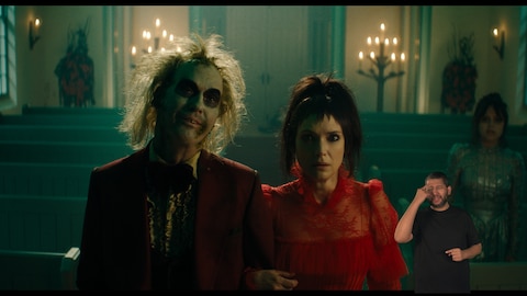 Beetlejuice e Lydia Deetz estão de braços dados dentro de uma igreja iluminada por velas, enquanto Astrid Deetz observa ao fundo. Beetlejuice veste um terno vermelho surrado, e Lydia usa um vestido vermelho formal. O intérprete de Libras, Andrey Baptista, está no canto inferior direito e sinaliza "pensar".
