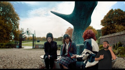 Lydia Deetz, Astrid Deetz e Delia Deetz têm uma conversa enquanto estão sentadas em um banco ao redor de uma grande escultura. O intérprete de Libras, Andrey Baptista, está no canto inferior direito e sinaliza "lágrimas".