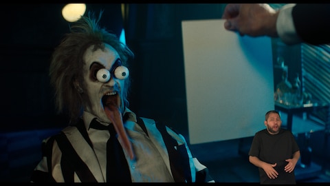 Beetlejuice grita de forma caricatural com os olhos esbugalhados, a boca escancarada e uma língua absurdamente longa para fora. O intérprete de Libras, Andrey Baptista, está no canto inferior direito e sinaliza "gritando".