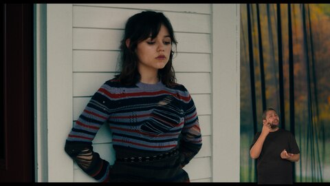 Astrid Deetz se apoia em um edifício de tábuas brancas com o olhar baixo.O intérprete de Libras, Andrey Baptista, está no canto inferior direito e sinaliza "ver".