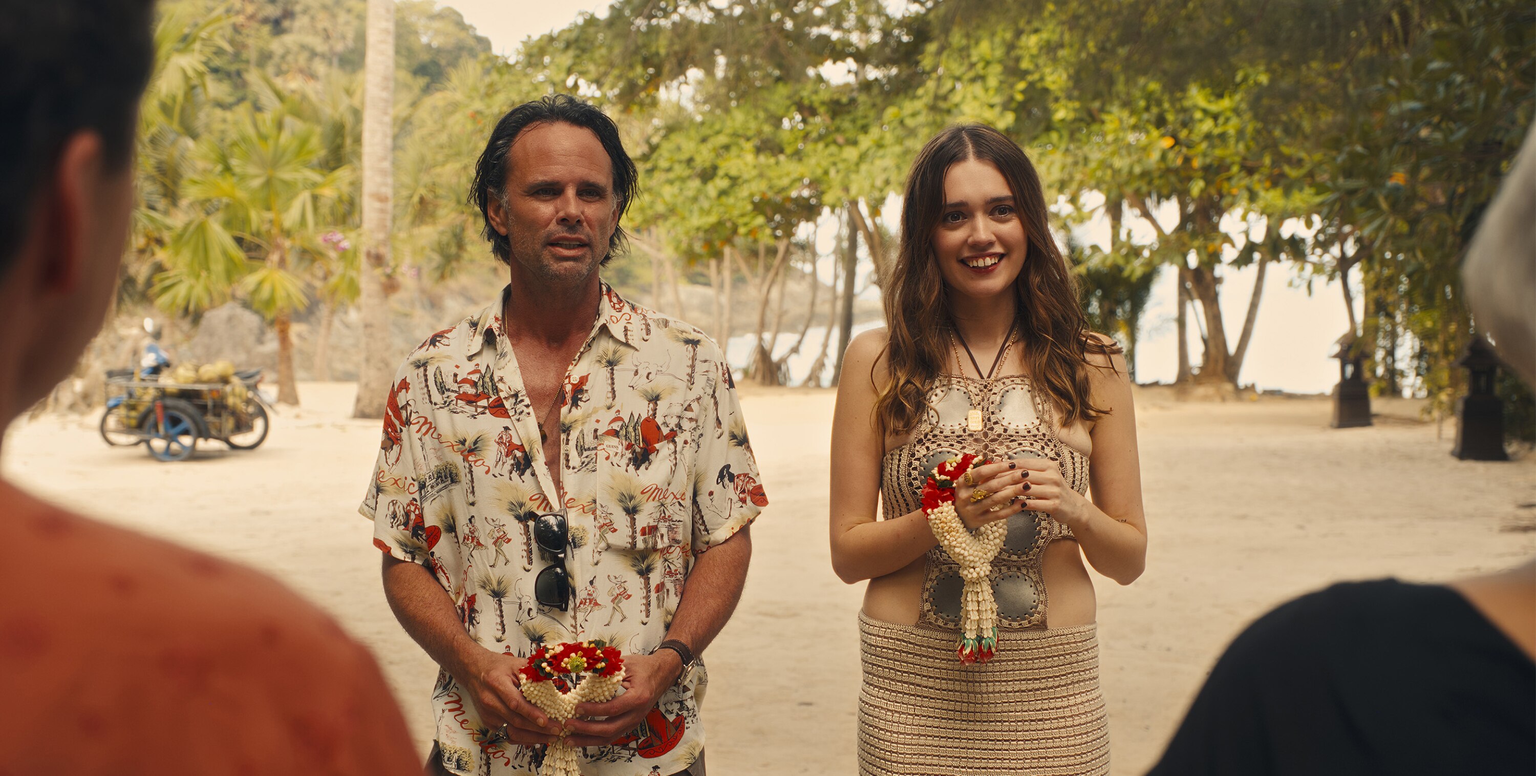 Rick Hatchett (Walton Goggins) and Chelsea (Amy Lou Wood) stand side by side, holding Thai flower garlands and looking expectedly at two people who have their backs to the camera. 