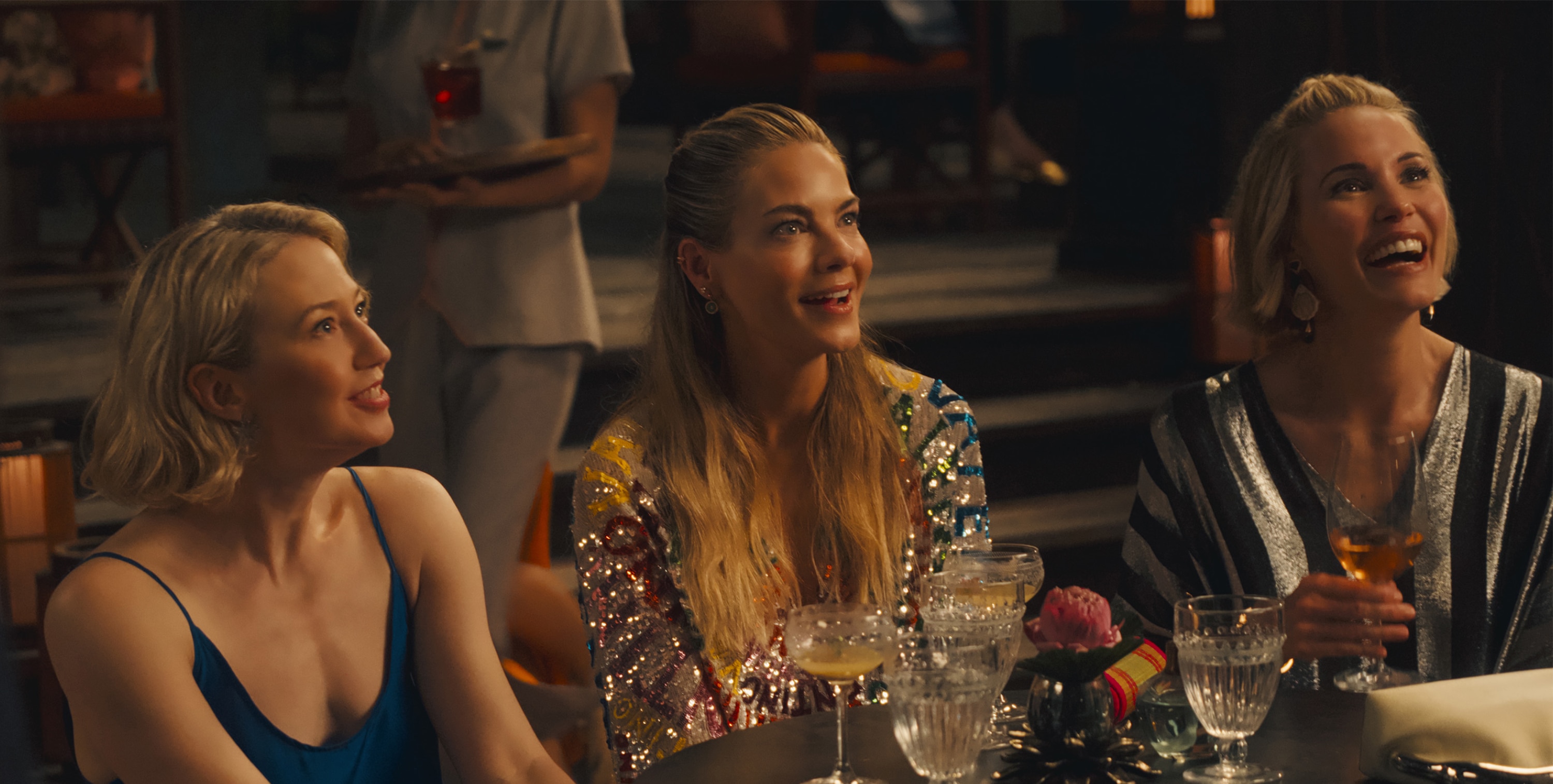 Laurie (Carrie Coon), Jaclyn (Michelle Monaghan), and Kate (Leslie Bibb) sit at a round table and smile at something out of frame.  