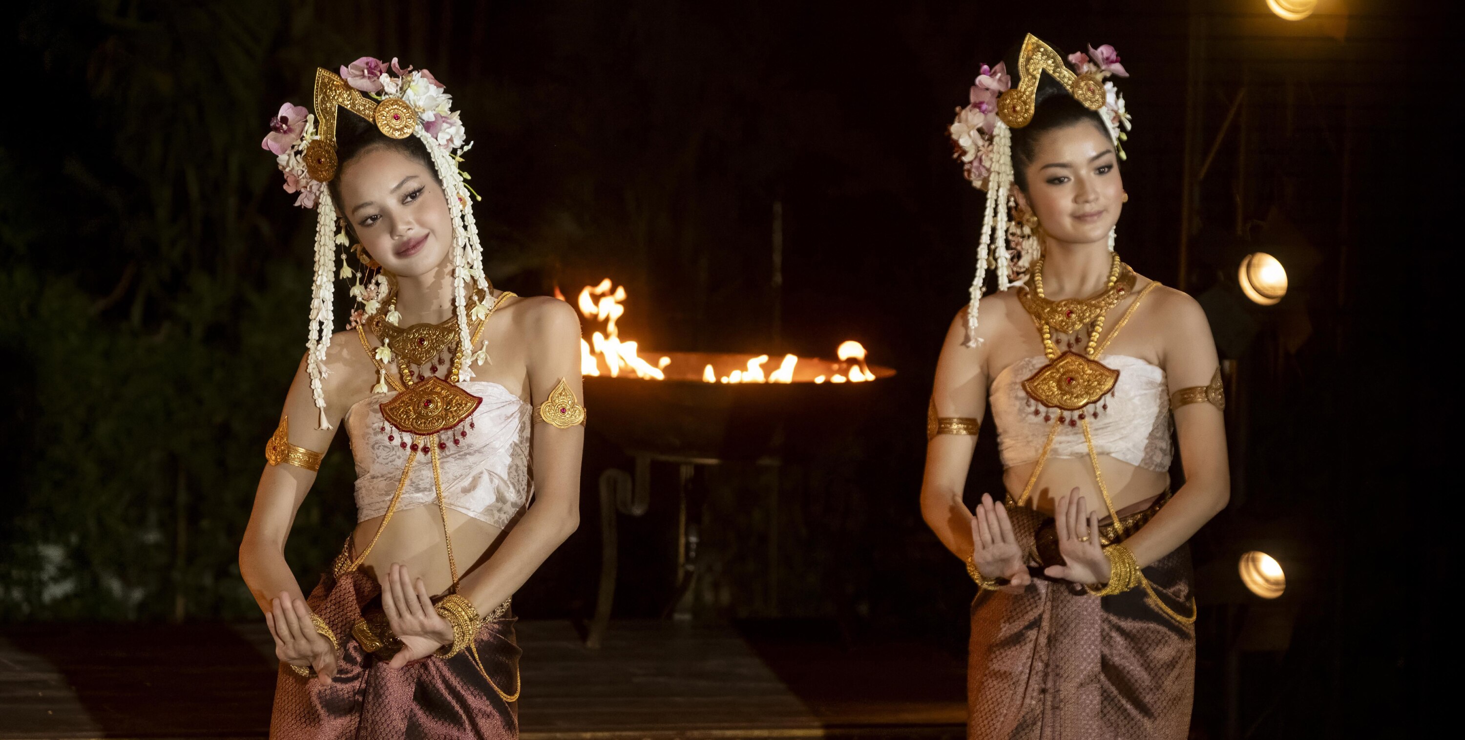 Mook (Lalisa Manobal) and another dancer perform a traditional Thai dance in ornate costumes and headpieces, with a fire pit glowing in the background. 