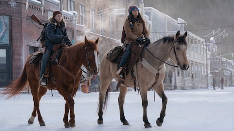 On the left, Ellie sits on a brown horse, her feet in the stirrups and hands holding the reins. She wears a blue coat, a knit hat, a pistol in a holster strapped to her right thigh, and a backpack with the handle of a long gun sticking out of the top. On the right, Dina sits on a light tan horse, her feet in the stirrups and hands holding the reins. She wears a brown coat, a knit hat, a knife and a pistol in holsters strapped to her right thigh, and a backpack. They both look at something out of frame.