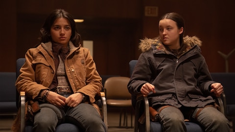 Dina and Ellie sit in chairs. Dina stares ahead while Ellie looks to her side at Dina.