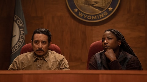 Tommy and Maria sit in high-back chairs covered in a red jacquard fabric and look at something out of frame. Maria rests her chin on her fist. A partially visible seal on the wall reads “Wyoming,” and a partially visible flag behind Tommy reads “Jackson.”