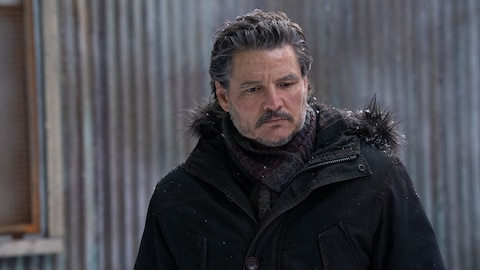 Joel stands outside. There is snow on his hair, eyebrows, beard, and hood of his black coat. He looks down at something out of frame.