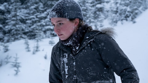 Abby stands outside in snow-covered woods. There is snow on her hat, sweater, and coat. She looks down at something out of frame.