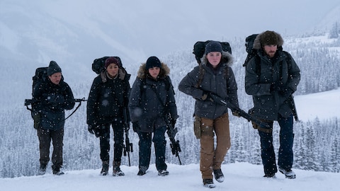 Abby and four other people walk through snow-covered woods. They all carry long guns.