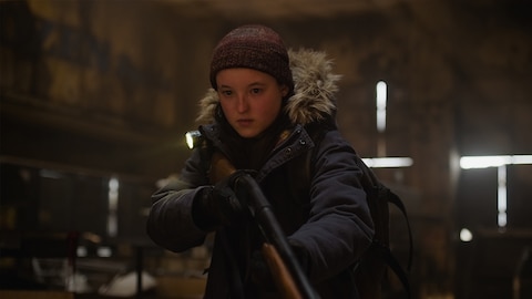 Ellie stands with her long gun in her hands, the barrel pointing down. A small flashlight attached to her shoulder is illuminated. She looks at something out of frame.