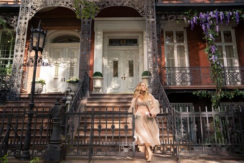 Carrie stands outside her new home. The entrance features two large white doors, with potted plants on each side and ornate black iron railings. She is wearing a long, flowing dress. 