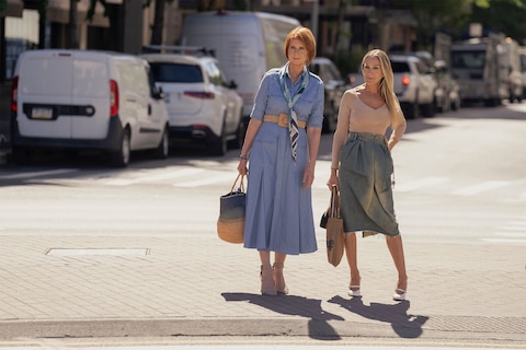 Miranda is wearing a light blue dress, and Carrie is wearing a beige top and green skirt. Both hold handbags as they meet on a New York City street.