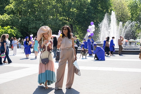Carrie and Seema enjoy ice cream in the park. In the background, a graduation celebration is taking place near a large fountain.