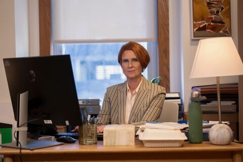 Miranda is seated at a desk, working on a computer. On the desk are a green water bottle, scattered papers, and various office supplies. 