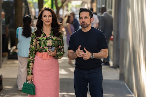 Charlotte and Anthony stroll together on the sidewalk. She is wearing a patterned blouse with a pink skirt and is carrying a green handbag. He is in a black T-shirt and dark jeans.