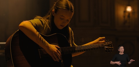 Ellie sits in a dimly lit room and tunes an acoustic guitar. ASL interpreter Leila Hanaumi signs “guitar playing.”