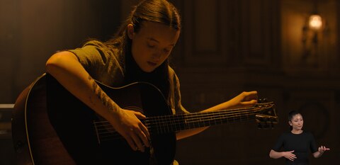 Ellie sits in a dimly lit room and tunes an acoustic guitar. ASL interpreter Leila Hanaumi signs “guitar playing.”