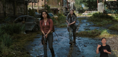 Dina and Ellie walk down the middle of a deserted city street with their handguns drawn and pointed down at the ground. ASL interpreter Leila Hanaumi signs “go ahead.”