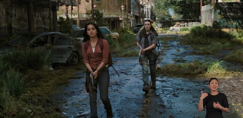 Dina and Ellie walk down the middle of a deserted city street with their handguns drawn and pointed down at the ground. ASL interpreter Leila Hanaumi signs “go ahead.”
