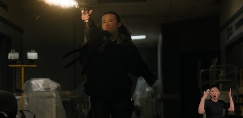 Ellie charges through a dimly lit room while firing her handgun. ASL interpreter Leila Hanaumi signs “intruder.”