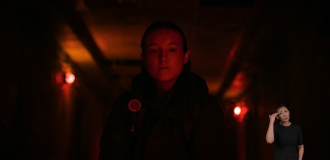 Ellie stands in a room bathed in red light and looks down at something out of frame. ASL interpreter Leila Hanaumi signs “I know.”