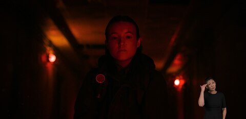 Ellie stands in a room bathed in red light and looks down at something out of frame. ASL interpreter Leila Hanaumi signs “I know.”