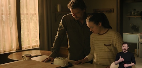 Joel and Ellie stand in a kitchen and look down at a small cake on a counter. ASL interpreter Daniel Durant signs “Happy Birthday.”
