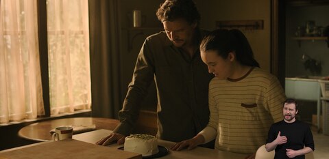 Joel and Ellie stand in a kitchen and look down at a small cake on a counter. ASL interpreter Daniel Durant signs “Happy Birthday.”