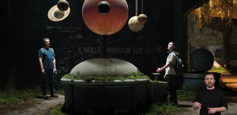 Joel and Ellie stand in an abandoned museum and look up at a large-scale model of the solar system. A mural to their left reads, “A Walk Through the Stars.” ASL interpreter Daniel Durant signs “pointing.”