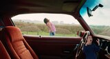 From inside the front seat of a classic car, we see Garrett standing outside the car with his head tilted back, yelling at the sky. ASL interpreter Sophia Morales signs “screaming.”