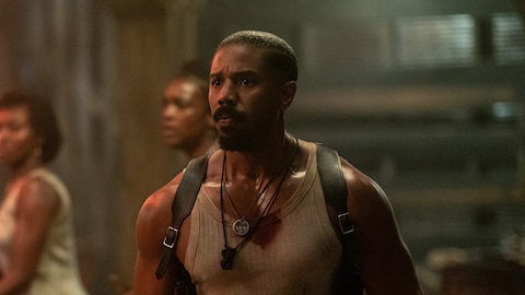 Smoke stands in the foreground in a bloodied tank top and wearing a double-draw handgun shoulder holster. Two people stand in the background. They all look at something out of frame. 