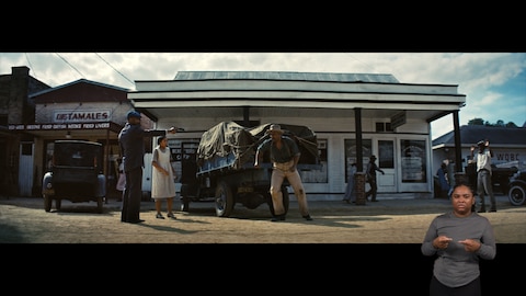 A row of businesses, including a restaurant, a cafe, and a radio station, lines the background. In the foreground, Smoke points a handgun at a man by a truck covered with a tarp. The man is trying to run away. A girl stands near Smoke with a shocked expression, and a man at the edge of the frame stands with his hands above his head in surrender. BASL interpreter Nakia Smith signs “hurt.”