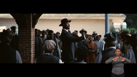 Stack stands in the middle of a crowd, slightly elevated. He points to something out of frame while speaking. BASL interpreter Nakia Smith signs “ready.”