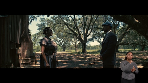 Annie and Smoke stand five feet apart and look at one another. They are shaded by the large trees surrounding them. BASL interpreter Nakia Smith signs “old.”
