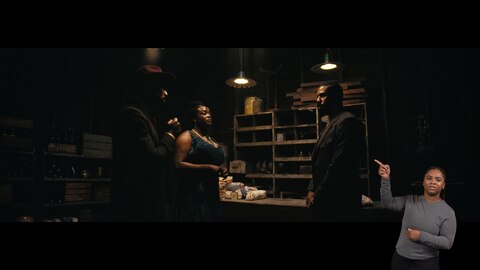 Stack, Annie, and Smoke stand in a dimly lit room and look at one another. Behind them is a mostly empty wood shelving unit and a counter with three packages of Wonder bread and other packaged goods. BASL interpreter Nakia Smith signs “point.”