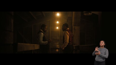Smoke and Stack stand in a dimly lit room. Smoke leans against a counter and looks down at his hands while smoking a cigarette. Stack stands two feet from Smoke and looks at him while talking. BASL interpreter Nakia Smith signs “total.”