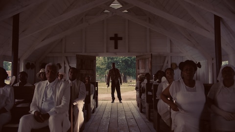 Sammie, wearing bloodied clothes, stands in the doorway of a church with a white-painted interior and stares straight ahead. The parishioners sit in pews on either side of him, all dressed in white. BASL interpreter Nakia Smith signs “let it shine.” 
