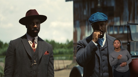Smoke and Stack stand outside next to one another while looking at something out of frame. X smokes a cigarette. BASL interpreter Nakia Smith signs “you.” 