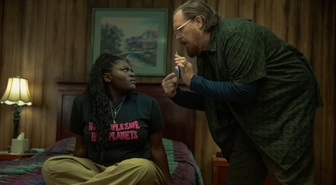 Leoto Adebayo sits on a bed with a multicolored quilt in a wood-paneled room. She is wearing a black T-shirt that reads “HOT PEOPLE SAVE HOT PLANETS.” John Economos stands beside her, leaning forward and pointing at his phone screen. A lamp and framed picture are in the background.
