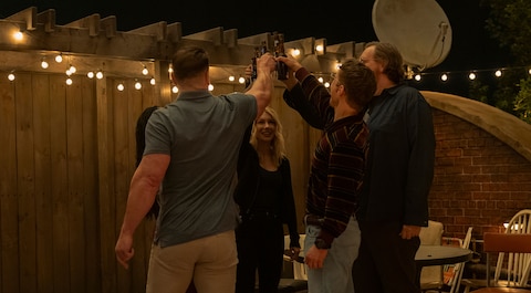 Christopher Smith, Leoto Adebayo, Emilia Harcourt, John Economos, and Adrian Chase stand outdoors at night. They raise their drinks in a toast beneath string lights. Tables and chairs surround them, with a wooden fence in the background and a large satellite dish to the right.