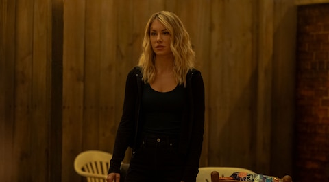 Emilia Harcourt stands in a dimly lit, wood-paneled room. She has a cut above her right eyebrow. Beside her is a round table with an ashtray, a mug, a set of keys, and a bag of chips. Two plastic chairs are behind her against the wall.