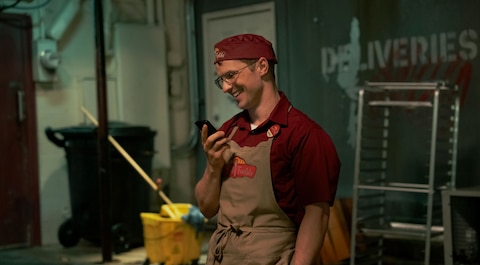 Adrian Chase, a busboy, stands in an alley wearing a red cap, red shirt, and beige apron. He holds a phone in one hand while smiling. Behind him are a trash bin, a bucket and mop, and a metal rack.