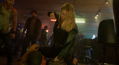 Emilia Harcourt stands over a man lying on the ground. Her fist is raised as if she is about to strike him. Two others watch from the background in a dimly lit bar with stools and tables.