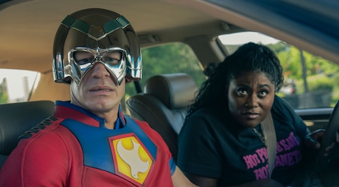 Peacemaker and Leoto Adebayo sit inside a car. She is in the driver’s seat. Both are looking out of the passenger side at something out of view.
