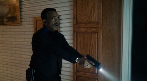 Langston Fleury points a flashlight-mounted gun downward inside a room with a brick wall.