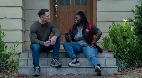 Christopher Smith and Leoto Adebayo sit on the front steps of a house with a wooden door. Green shrubs are on either side of the steps. She smiles as she holds a phone in her hand.