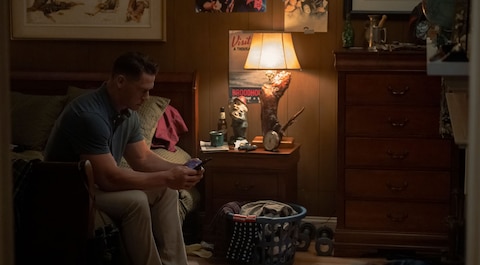 Christopher Smith sits on the edge of a bed in a dimly lit, wood-paneled room. He looks intently at his phone. Posters of musicians and bands hang on the wall. A lamp on the nightstand casts light over a cluttered nightstand, and a laundry basket filled with clothes is on the floor near the bed.